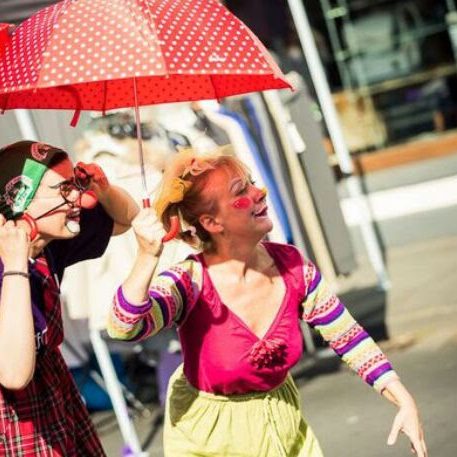 Straßentheater Zwei Clowns mit einem roten Regenschirm und bunter Kleidung, die fröhlich auftreten.